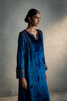 Woman wearing a blue dress with intricate patterns against a textured wall.