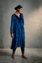 Woman wearing a blue embroidered traditional outfit against a gray wall.