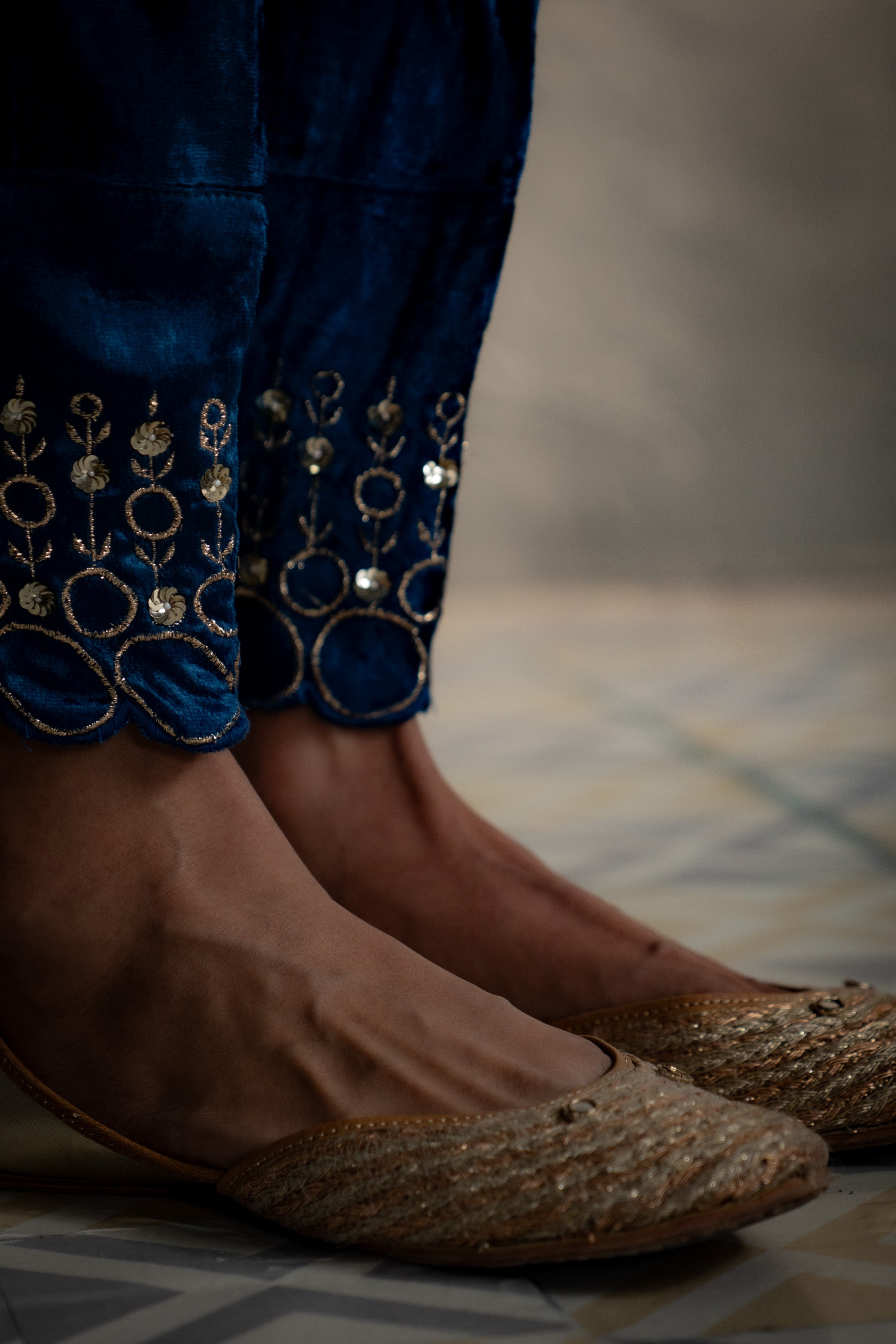 Close-up of feet wearing blue embroidered pants and gold sandals on a patterned floor.